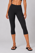 A woman modeling a matching black activewear set consisting of a sports bra and high-waisted capri leggings, standing against a light gray studio background.