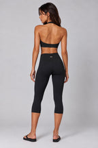 A woman is shown from the back wearing a matching black activewear set consisting of a halter-style sports bra and high-waisted capri leggings, paired with black flat sandals, against a light gray studio background.