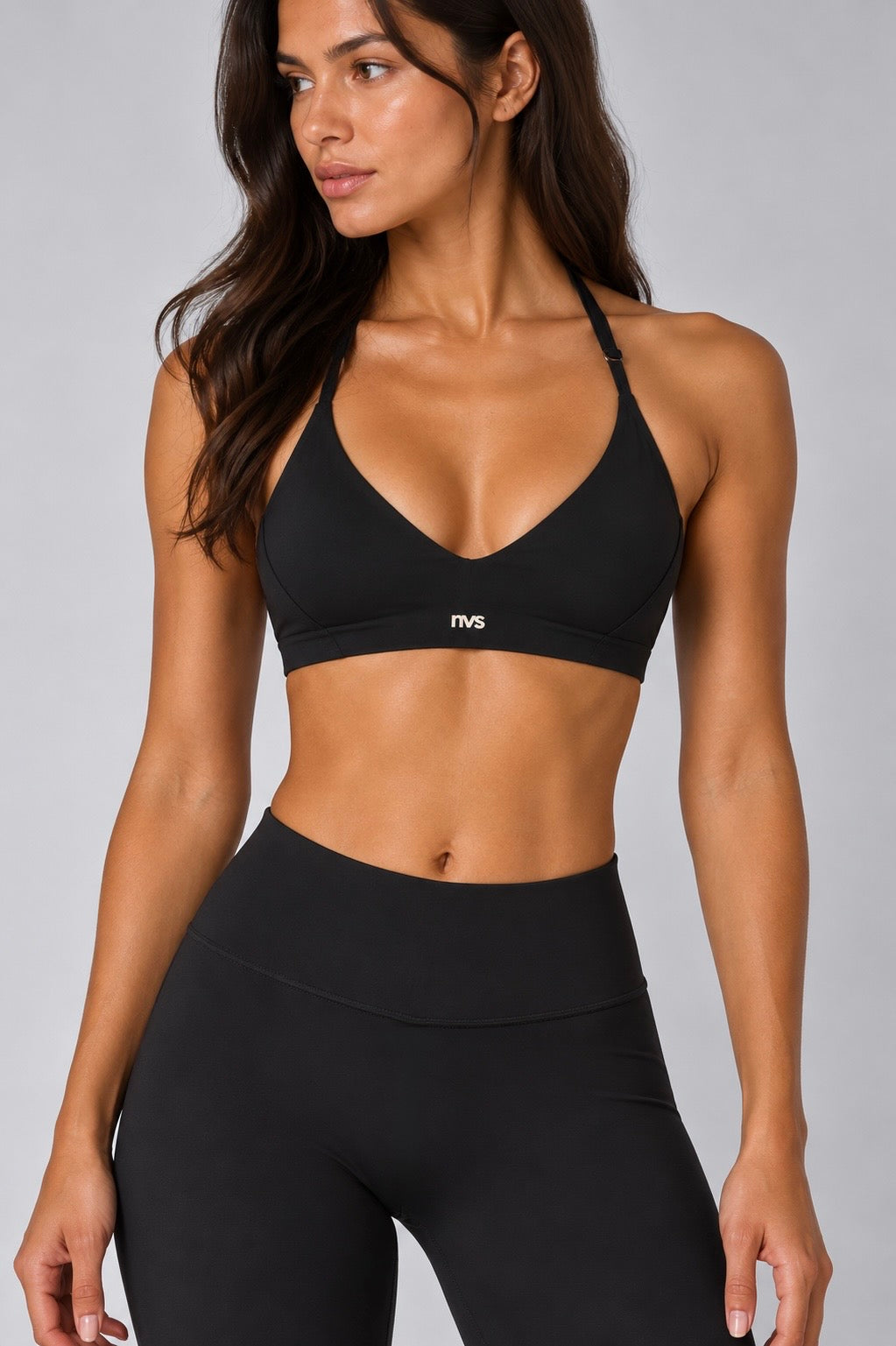 A woman modeling a black athletic sports bra and matching high-waisted leggings against a light gray studio background. The sports bra has a small white logo on the front band.