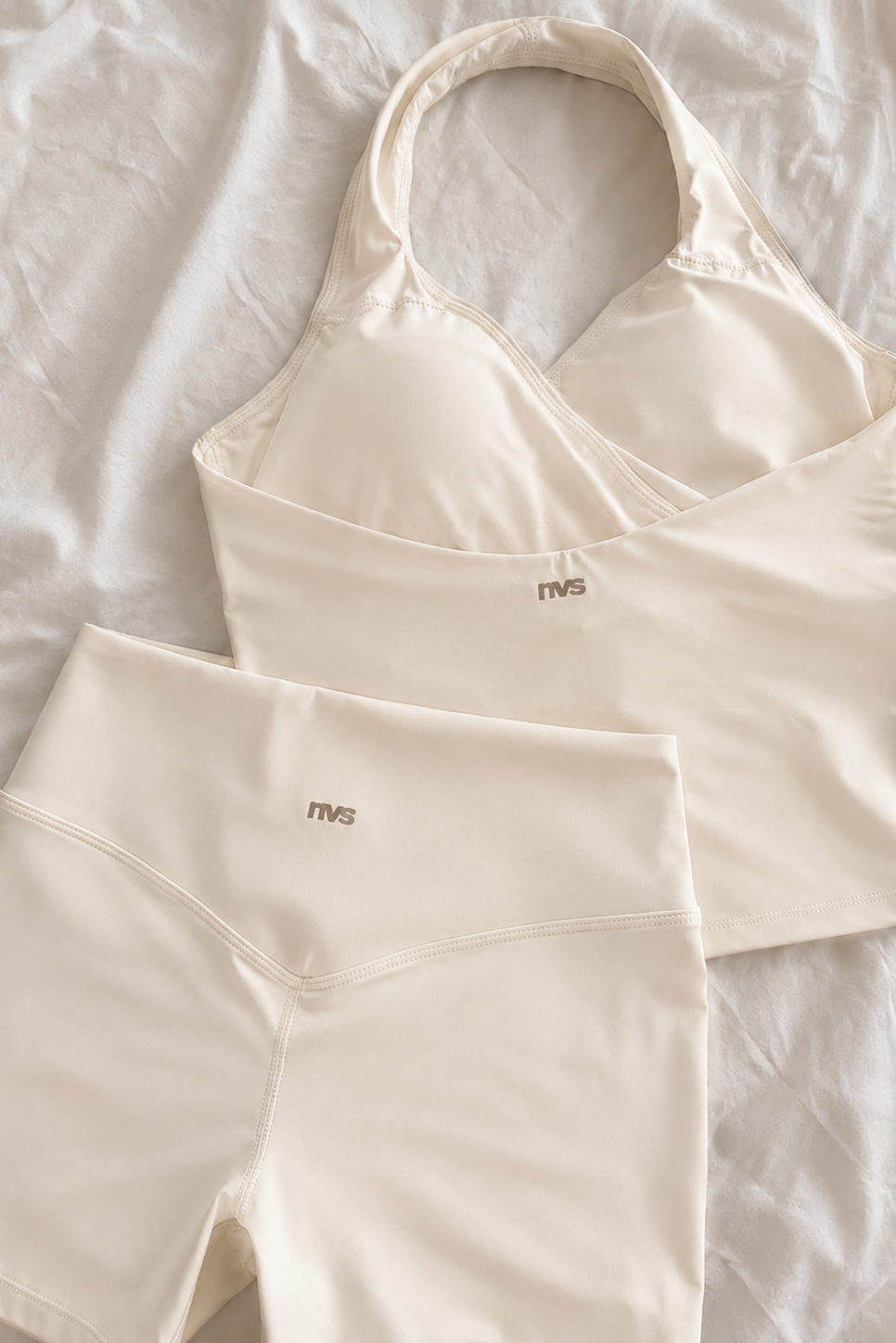 Cream-colored activewear set consisting of a halter-neck sports bra and matching high-waisted shorts, laid flat on a white textured fabric background. The brand name 'mvs' is printed in small brown text on the front of both garments.