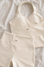 Cream-colored activewear set consisting of a halter-neck sports bra and matching high-waisted shorts, laid flat on a white textured fabric background. The brand name 'mvs' is printed in small brown text on the front of both garments.