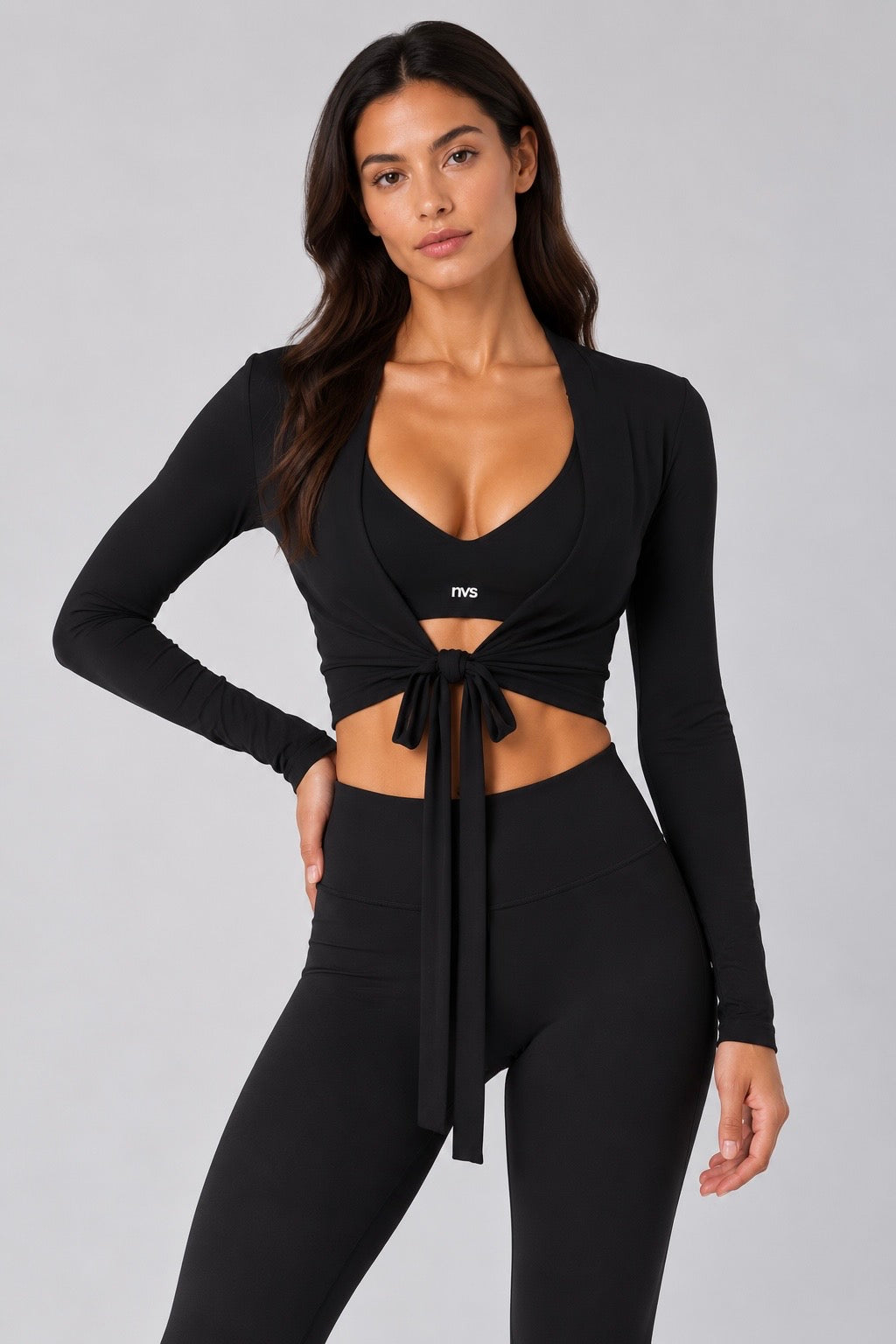 A woman modeling a black long-sleeve wrap-style crop top with a front tie detail and matching black high-waisted leggings against a light gray studio background.