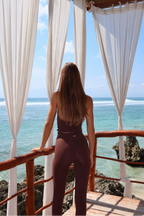 A woman stands on a wooden deck overlooking the ocean, viewed from behind. She is wearing a matching espresso brow colour activewear set consisting of a haltertop and leggings.